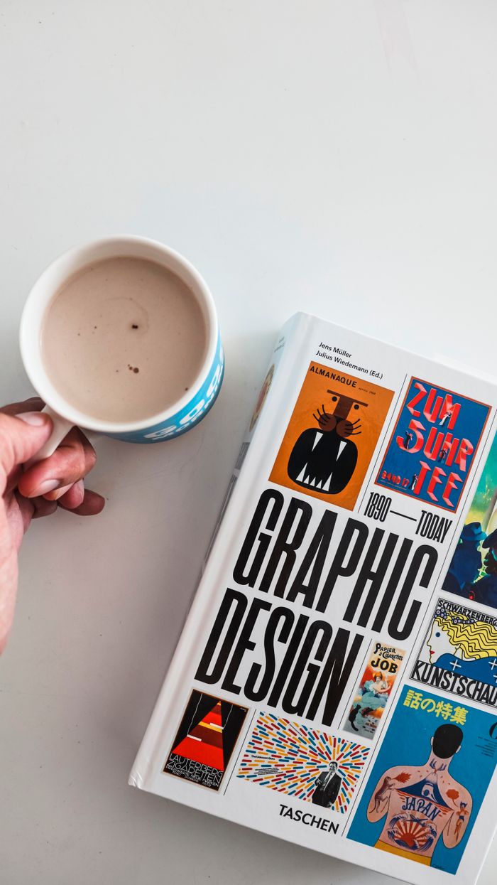 Services Close-up of a hand holding a coffee cup next to a graphic design book on a white surface.